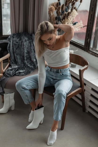 Schöne junge stilvolle blonde Frau in einem weißen T-Shirt mit Jeans sitzt in einem Sessel und zieht weiße Schuhe in einem Raum in der Nähe der Fenster an