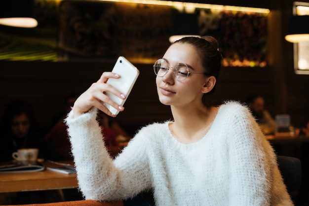 Schöne junge Studentin in weißem Pullover und Brille macht Selfie auf ihrem Smartphone sitzt in einem gemütlichen Café