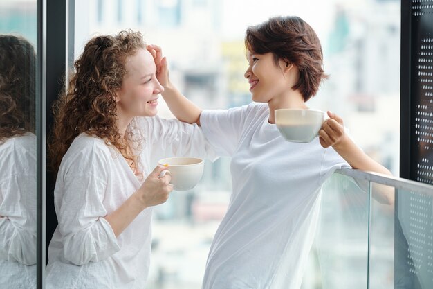 Schöne junge Vietnamesin, die Haare ihrer Freundin berührt, wenn sie Morgenkaffee auf dem Balkon trinken