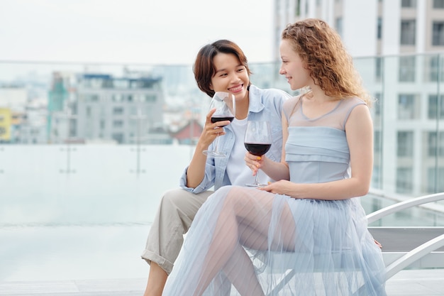 Schöne junge vietnamesische Frau in der Liebe, die Glas Rotwein trinkt und ihre lächelnde Freundin ansieht