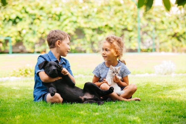 Schöne Jungen und Mädchen plaudern glücklich miteinander, sitzen auf Gras und umarmen ihre Haustiere Sommerferien im Freien mit Haustieren Große Wiese und Bäume im Hintergrund