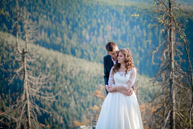 Schöne Jungvermählten, die gegen die Berge umarmen. Stilvolle Braut und schöne Braut stehen auf der Klippe. Hochzeitsporträt. Familienfoto