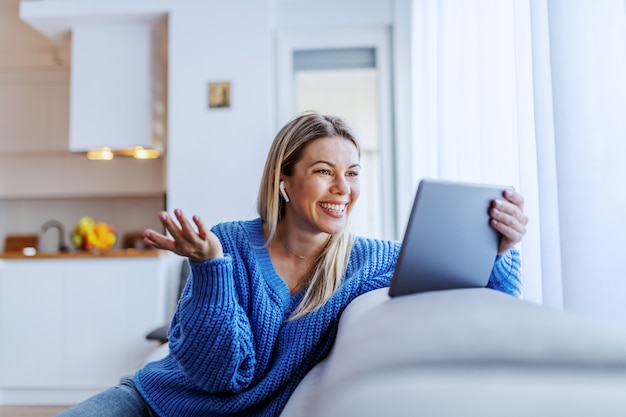 Schöne kaukasische blonde Frau gekleidet im blauen Pullover, der auf Sofa im Wohnzimmer sitzt und Videoanruf über Tablette hat.