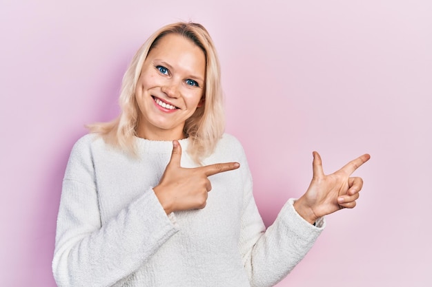 Schöne kaukasische blonde Frau trägt einen lässigen Winterpullover, lächelt und blickt in die Kamera, während sie mit zwei Händen und Fingern zur Seite zeigt.