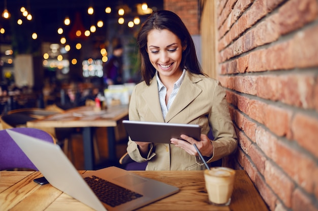 Schöne kaukasische Brünette gekleidet intelligentes lässiges Sitzen im Café und unter Verwendung der Tablette. Auf dem Schreibtisch stehen Laptop und Kaffee.