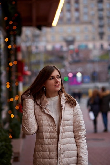 Schöne kaukasische Frau auf der Straße in der Nähe von geschmückter Vitrine Stilvolle Freizeitkleidung für den Winter Festliche Stadt Abendlichter in der Stadt