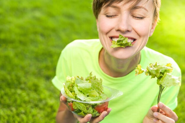 Schöne kaukasische Frau, die Salat über grünem natürlichem Hintergrund isst