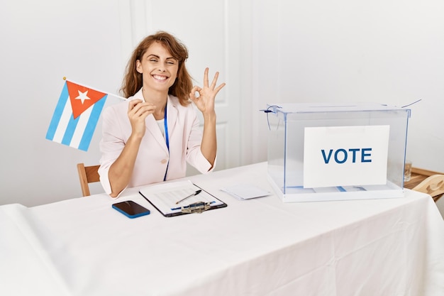 Foto schöne kaukasische frau im politischen wahlkampf, die die kubanische flagge in der hand hält und mit den fingern ein „ok“-zeichen macht, freundlich lächelt und ein ausgezeichnetes symbol zeigt