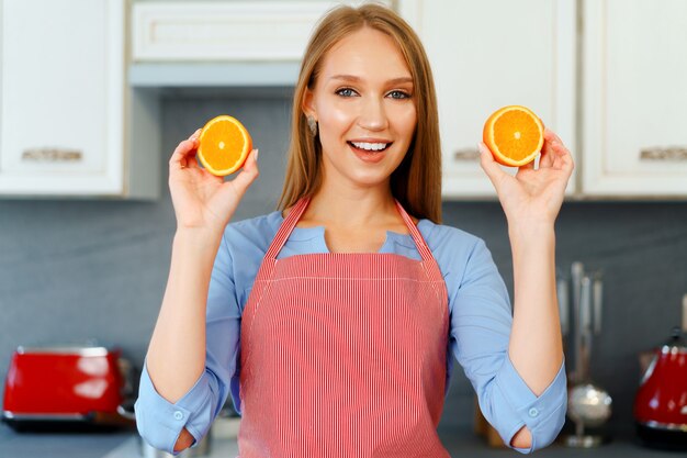 Schöne kaukasische Frau in der roten Schürze, die reife Orangen beim Stehen in der Küche hält