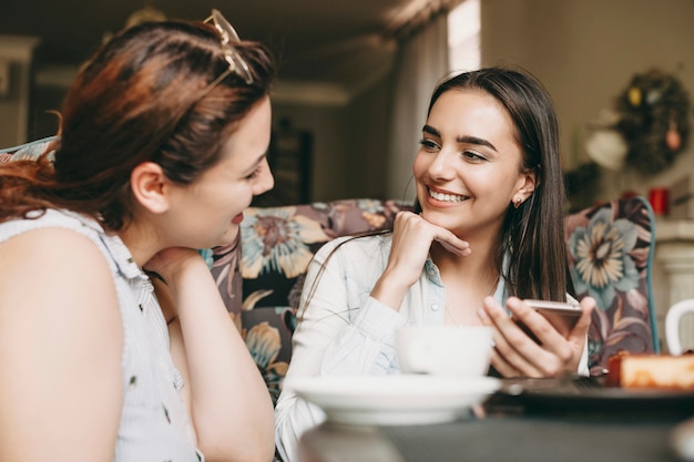 Schöne kaukasische Frau mit dunklen langen Haaren, die in einem lächelnden Kaffeehaus sitzen, während sie ihre Freundin mit einem Smartphone in ihren Händen hören.