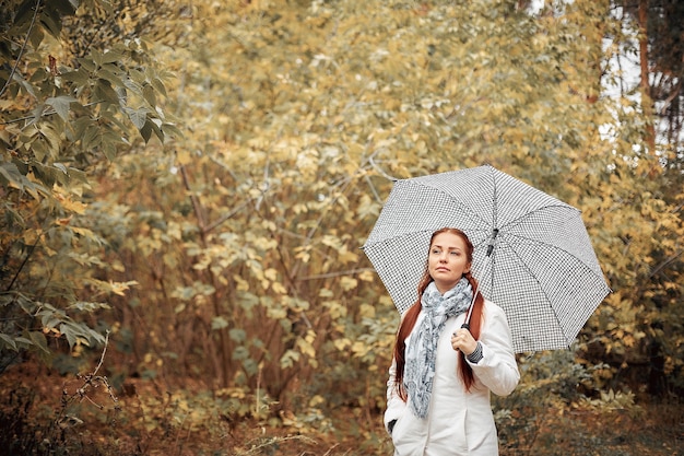 Schöne kaukasische Frau mittleren Alters mit roten Haaren mit einem Regenschirm im Park an einem wolkigen Herbsttag.