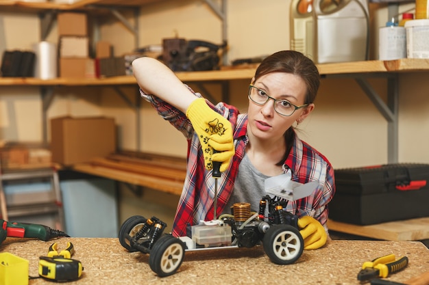 Schöne kaukasische junge braunhaarige Frau in kariertem Hemd, grauem T-Shirt, gelben Handschuhen, die Spielzeugauto-Eisenmodellkonstrukteur machen, in der Tischlerei am Holztischplatz mit verschiedenen Werkzeugen arbeiten.