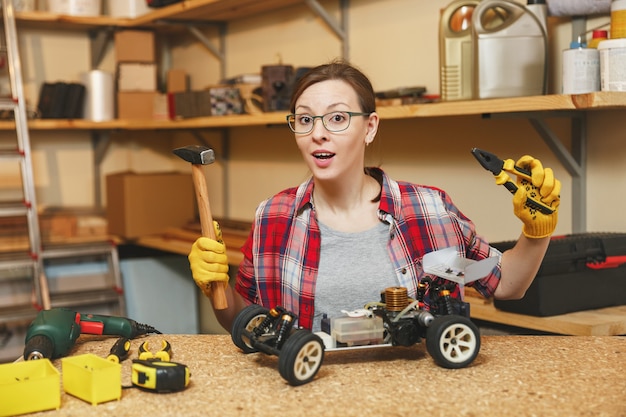 Schöne kaukasische junge braunhaarige Frau in kariertem Hemd, grauem T-Shirt, gelben Handschuhen, die Spielzeugauto-Eisenmodellkonstrukteur machen, in der Tischlerei am Holztischplatz mit verschiedenen Werkzeugen arbeiten.