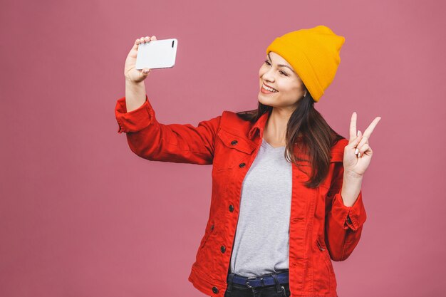 Schöne kaukasische junge Frau, die Handy hält und für selfie aufwirft, mit flirty Lächeln schauend. Attraktive Frau, die Videoanruf macht. Isoliert über rosa Wand.