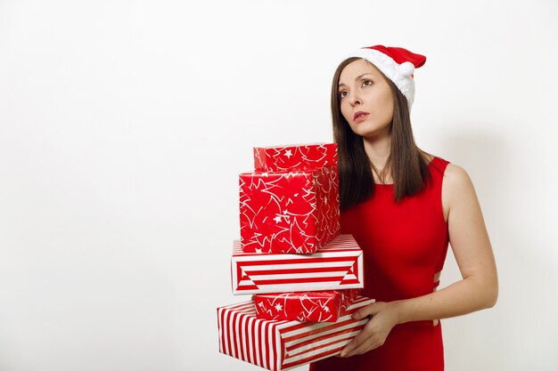 Schöne kaukasische junge verärgerte frustrierte und traurige Frau, die im roten Kleid und im Weihnachtshut gekleidet wird, der Geschenkboxen auf weißem Hintergrund hält. Sankt-Mädchen mit dem Geschenk lokalisiert. Neujahrsfeiertag 2018 Konzept.