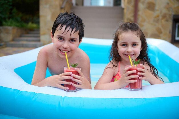 Schöne kaukasische Kinder, Junge und Mädchen, die gesunden, frisch gepressten Fruchtcocktail trinken, während sie Spaß in einem Wasserbecken haben