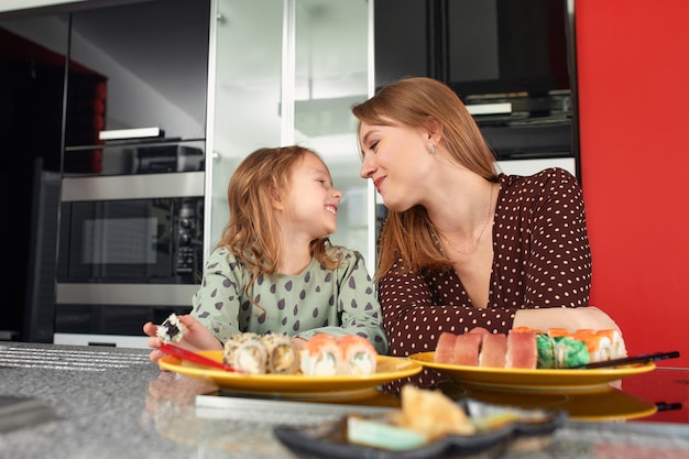 Schöne kaukasische Mutter und Tochter, die Brötchen und Sushi beim Mittagessen mit der Familie zu Hause essen Lieferkost Traditionelles japanisches Essen