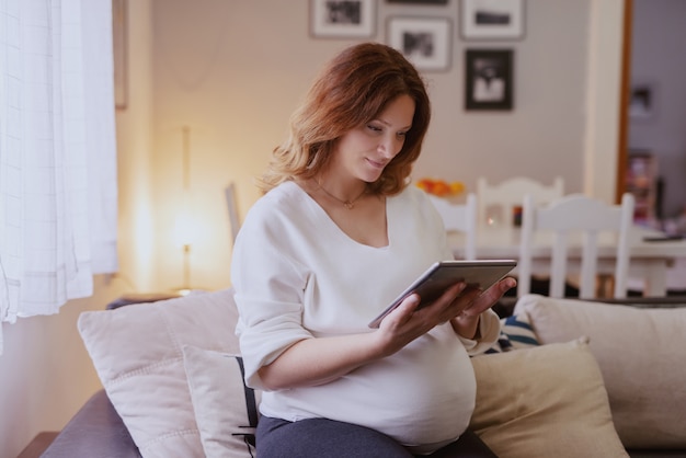 Schöne kaukasische schwangere Frau, die im Wohnzimmer im Sofa sitzt und Tablette verwendet.