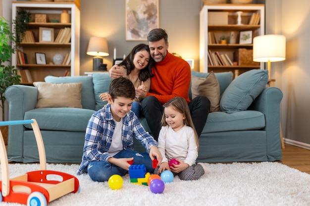 Schöne Kinder spielen, während die Eltern zusammen auf dem Sofa zu Hause entspannen, lächelnd, aktiver Junge unterhält sich mit einem Spielzeugwagen in der Nähe seiner Schwester auf dem Boden, glückliche Familie verbringt am Wochenende Zeit zusammen im Wohnzimmer.