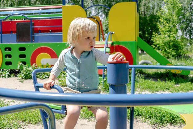 Schöne kleine blonde junge Kleinkind-Kind-Kind-Junge, der auf einer Schaukel, einem Kreisverkehr, einem Karussell im Spielplatzpark fährt Kinder körperliche emotionale Entwicklung Kindheit Kindertagesstätte Kindergartenkonzept