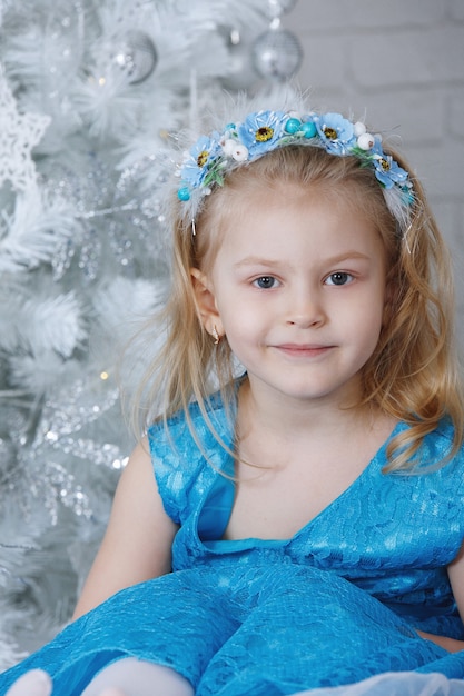 Schöne kleine Blondine, die auf dem Bett sitzt. In ihren Händen eine Weihnachtskugel. Das Mädchen im blauen Kleid.
