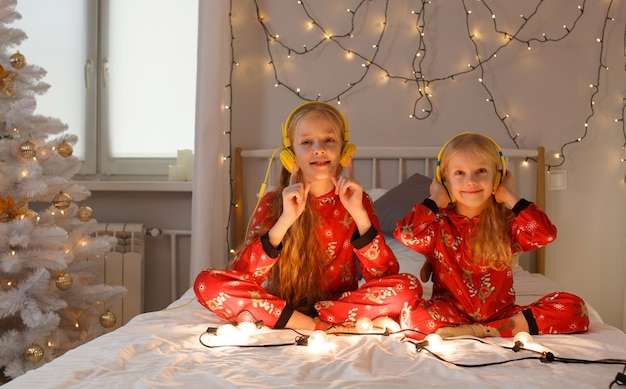 Schöne kleine Mädchen, die im Bett in hellen Weihnachtspyjamas sitzen und Musik mit Kopfhörern hören, heller heller Raum. Weihnachten. Neujahr