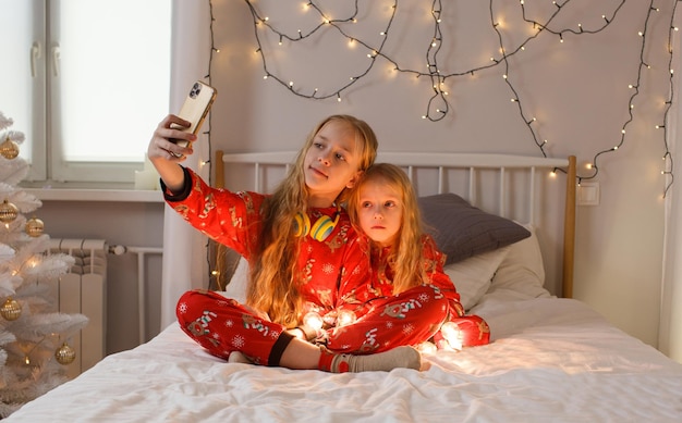 Schöne kleine Mädchen sitzen im Bett im hellen Weihnachtspyjama und machen Fotos von sich selbst am Telefon, ein heller Raum. Weihnachten. Neujahr