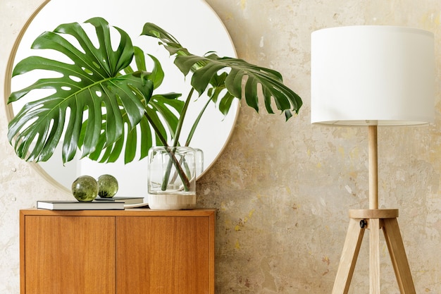 Schöne Komposition im modernen Retro-Interieur mit Design-Kommode, tropischem Blatt in Vase, rundem Spiegel, Lampe, Buch, Grunge-Wand und eleganten Accessoires in stilvoller Wohnkultur.