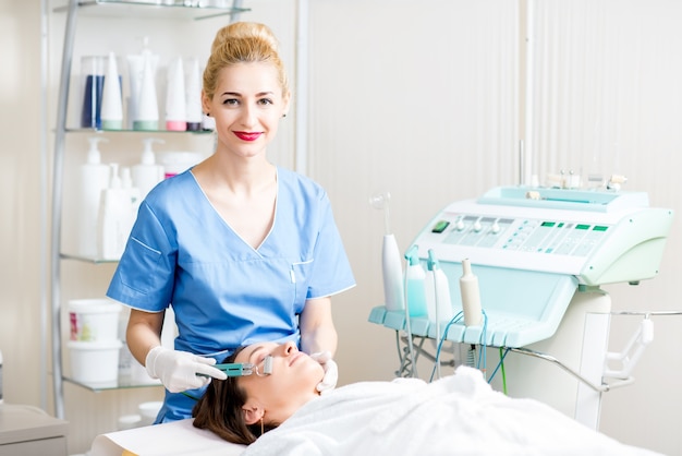 Schöne Kosmetikerin in Uniform, die dem Patienten im Kosmetikbüro eine Gesichtsbehandlung macht