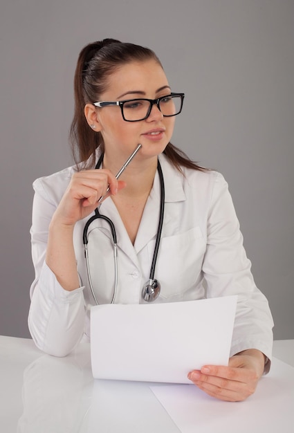 Schöne Krankenschwester in Robe und mit Stethoskop am Hals macht ihren Bericht am weißen Tisch