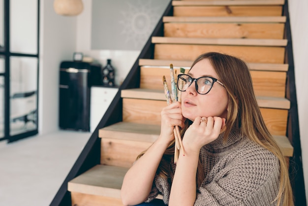 schöne Künstlerin in Brille und Freizeitkleidung, die mit Bürsten auf Treppen sitzt