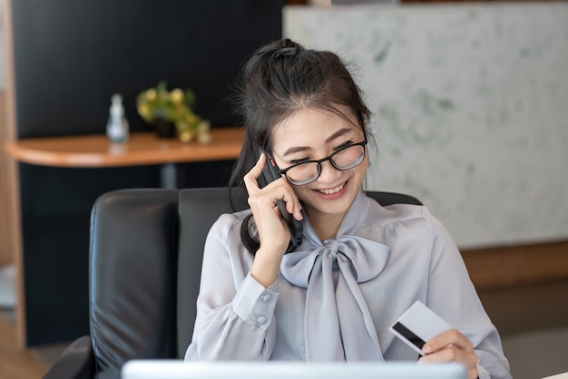 Schöne lächelnde asiatische Frau, die auf Handy glücklich hält Kreditkarte im Büro hält.