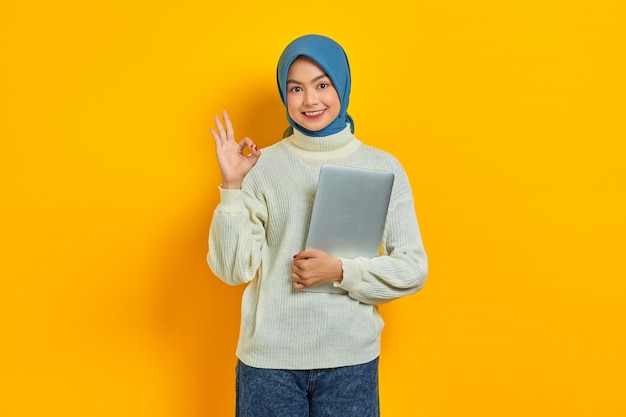 Schöne lächelnde asiatische Frau im weißen Pullover, die einen Laptop hält und eine Okay-Geste zeigt, demonstriert ein Symbol der Zustimmung, das über gelbem Hintergrund isoliert ist. Menschen religiöses Lebensstilkonzept