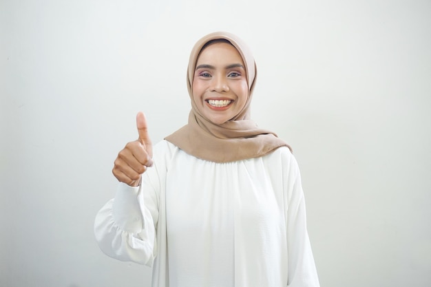 Schöne lächelnde asiatische Frau in weißer Freizeitkleidung, die Daumen nach oben zeigt, Geste mit der Hand