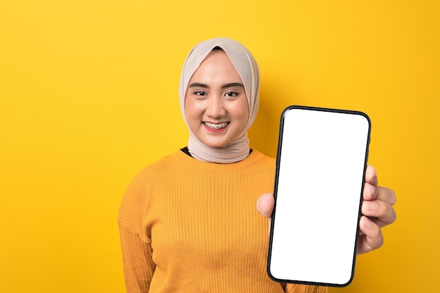 Schöne lächelnde asiatische Frau mit Hijab hält ein Smartphone mit einem leeren weißen Bildschirm, das auf einem gelben Hintergrund-Mockup isoliert ist