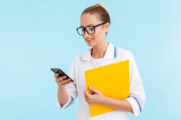 Schöne lächelnde blonde Ärztin in Uniform, die isoliert über blauer Wand steht, mit Handy, Notizblock hält