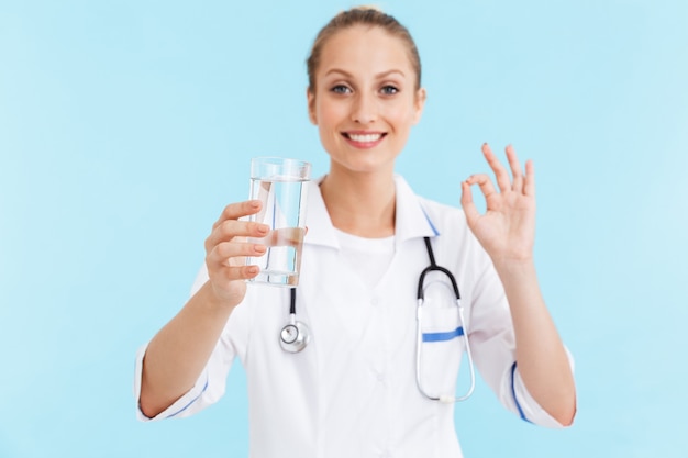 Schöne lächelnde blonde Ärztin in Uniform, die isoliert über blauer Wand steht und ein Glas Wasser zeigt, ok Geste