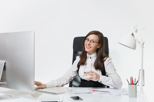 Schöne lächelnde braunhaarige Geschäftsfrau in Anzug und Brille, die mit einem Glas reinem Wasser am Schreibtisch sitzt und am Computer mit modernem Monitor mit Dokumenten im hellen Büro arbeitet working