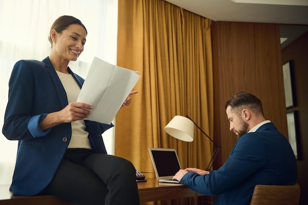 Schöne lächelnde, erfolgreiche, begeisterte Frau mittleren Alters in legerem Anzug, die auf einem Tisch mit Dokumenten sitzt, während ihr Partner geschäftlich auf der Laptop-Tastatur tippt, die im Hotelzimmer zusammenarbeitet?