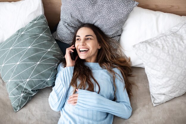 Foto schöne lächelnde frau, die auf handy spricht, während sie im gemütlichen bett zu hause liegt. menschen-, technologie- und kommunikationskonzept.