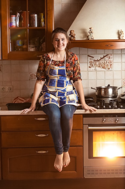 Schöne lächelnde Frau, die beim Backen im Ofen auf Tischplatte sitzt