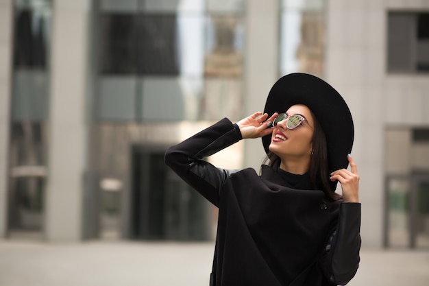 Schöne lächelnde Frau in schwarzem Hut mit breiter Krempe und Spiegelsonnenbrille