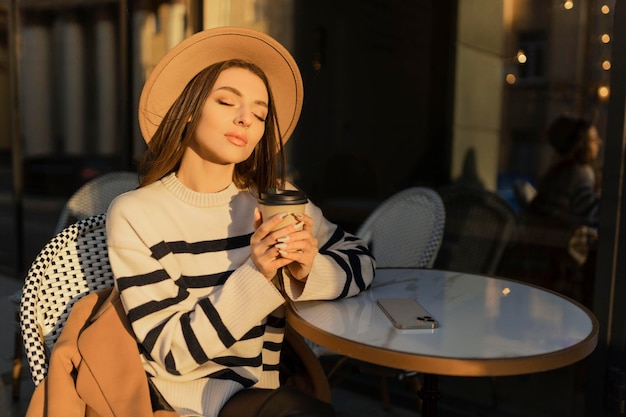 Schöne lächelnde Frau in stilvollem Outfit, die am Tisch in einem Hut und Pullover sitzt, romantische, fröhliche Stimmung, die auf einen Freund bei einem Date in einem Café wartet, Frühlingssommer-Modetrend, der Kaffee trinkt