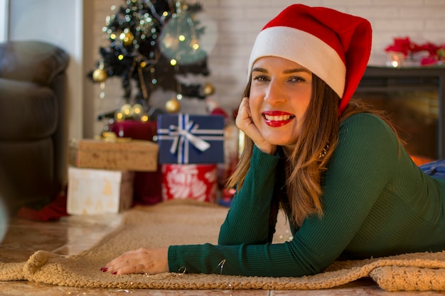 Schöne lächelnde Frau mit dem Weihnachtshut, liegend auf ihrem Teppich nahe bei Weihnachtsbaum und Geschenken