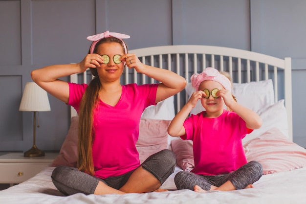 Schöne lächelnde Frau und ihre Tochter, die sich zu Hause amüsieren, indem sie Gurken wie Brillen vor ihre Augen halten