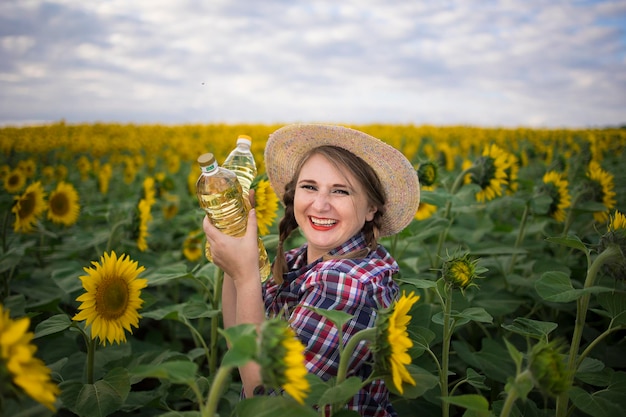 Schöne lächelnde, fröhliche Bäuerin mittleren Alters, die an einem sonnigen Tag Flaschen mit goldenem Sonnenblumenöl in ihren Händen in einem Erntefeld von Sonnenblumen hält