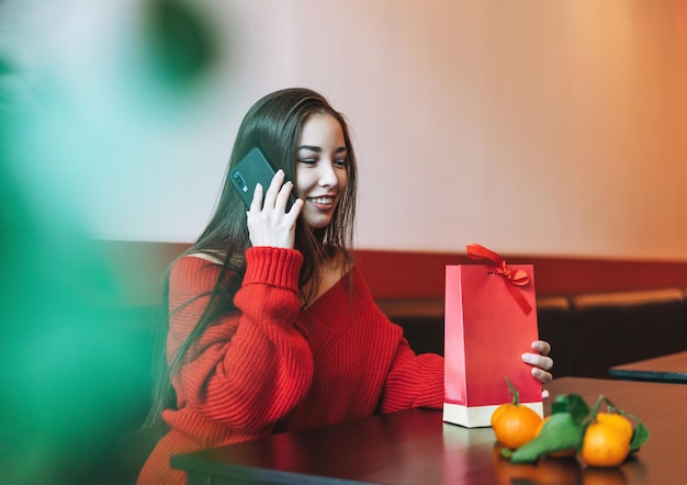 Schöne lächelnde junge Asiatin in Rot kleidet sich mit Handy mit Geschenktüte mit Mandarinen im Café