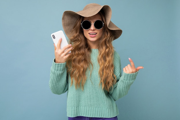 Schöne lächelnde junge blonde Frau mit Sonnenbrille und Hut isoliert auf dem Hintergrund mit Kopienraum, der Smartphone an der Kamera hält.