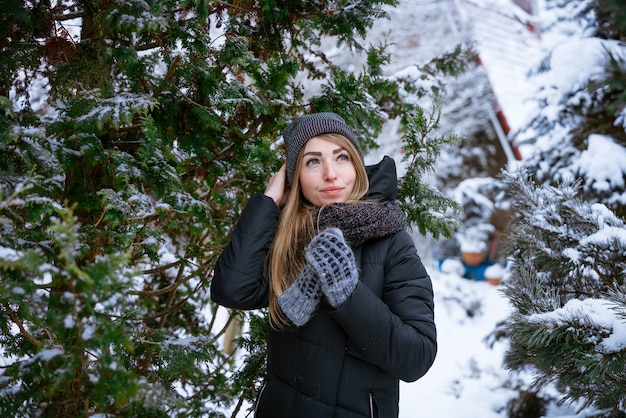 Schöne lächelnde junge Frau im Winter draußen in warmer Jacke genießt das Wetter unter den schneebedeckten...