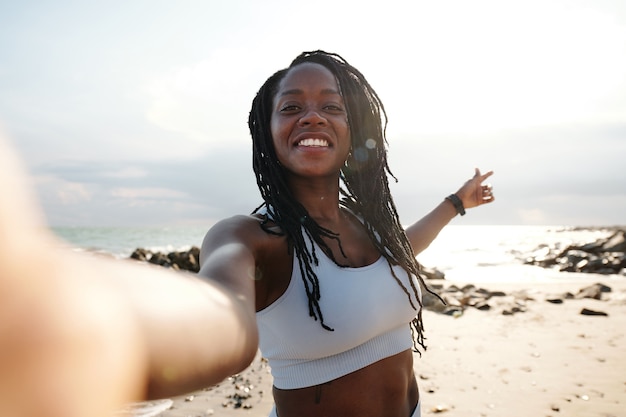 Schöne lächelnde junge Frau mit Dreadlocks, die nach dem Training am Morgen Selfie am Strand macht